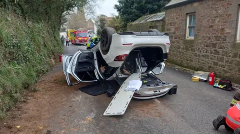 BBC A white car flipped onto its roof on a main road, with a fire engine and police car behind and people observing on the street. Brown debris is on the road, with a brick house on the right and trees and grass mounds on the left. A church can be seen in the distance.