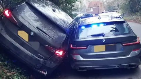 Unmarked police car on road to right with blue lights visible in back window. Alongside it is another black BMW vehicle leaning at an angle against the other car as it is up a grass verge - a brick house can be seen in the distance down the road.