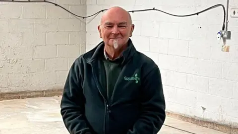 A man wearing a dark-coloured jacket and jeans stands in an empty unit with a metal door and white brick walls.