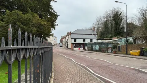 A view of the High Street in St Asaph, there are black railings and a large green tree on the left, cobbled pavements, a red tarmac road with zigzag road markings and white buildings with slate roofs in the distance