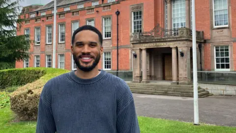 BBC/@lamarmangal Lamar in the foreground wearing a dark long-sleeved sweater and grey trousers, standing on a grass area with hands in pockets. Behind, a red-brick building labelled “Trafford Town Hall” has steps, columns, and large windows. Two tall flagpoles stand in front, one flying a Union flag. Low hedges border the grass, with a paved path leading to the entrance. The sky is overcast, and no bins or bags are visible.
