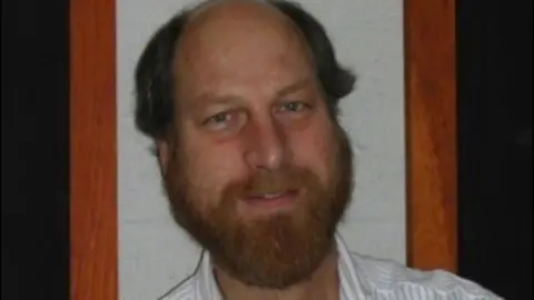 Carl Grillmair is seen smiling for a photo. He has a beard and is wearing a collared shirt 