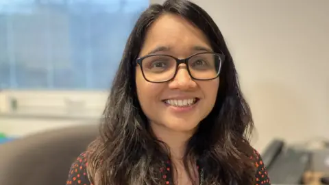 Frome GP Neha Bhagi smiles into the camera. She has long dark hair and glasses.