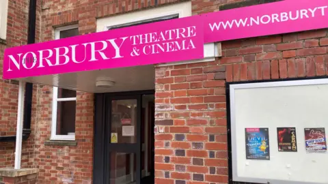 A picture of the outside of the Norbury Theatre in Droitwich, with a bricked frontage and a display cabinet showing images of upcoming performances.