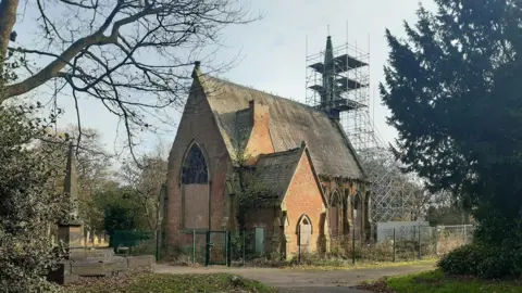 A dilapidated chapel surrounded by a metal fence. The building has boarded-up windows as well as scaffolding supporting an area at the back.