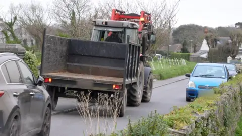 An image showing a road with a wall on one side and a pavement and gardens on the other side. There is a tractor with a trailer travelling on the left-hand side of the road facing away from the camera with its left-hand wheels on the pavement. Part of a black car is visible behind the tractor on the same side of the road. On the other side of the road a small blue car is in line with the front of the tractor and there is a second car behind it.  