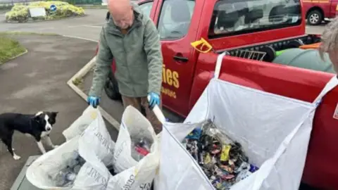 A man in a green coat with large white bags filled with rubbish. He looks down at them, there is a red van to the right and a black and white collie dog to the left. 