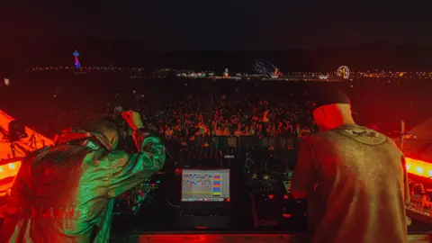 Jessie Myers A view looking out behind two DJs, with a laptop in front of them, to the crowd beyond at night.