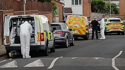 A road with three police vehicle and a silver car in the middle of them. The police vehicle nearest to the camera has its doors open as a forensic inspector looks inside