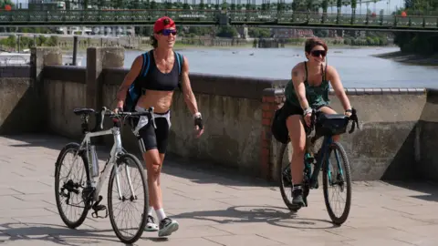 PA Media Two women in exercise clothing cycling along the River Thames in the sunshine. One is walking alongside their bike while the other rides theirs