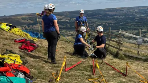 Edale Mountain Rescue Team Five rescuers sitting on grass wearing helmets and harnesses. They are surrounded by equipment. 