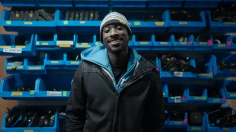 All Set West A young man wearing a blue hoodie underneath a black jacket. He has a striped beanie hat on and is standing in front of a wall of blue shelving units filled with filming equipment like batteries.