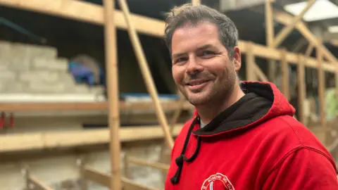 An unshaven man with dark hair wearing a red hoodie smiles at camera in front of a pile of wood in a barn.