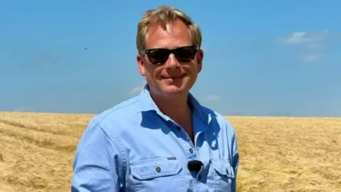 James Peck has short blond hair and is wearing black sunglasses and a blue shirt. He is standing in a field of yellow crops, and the sky is blue.