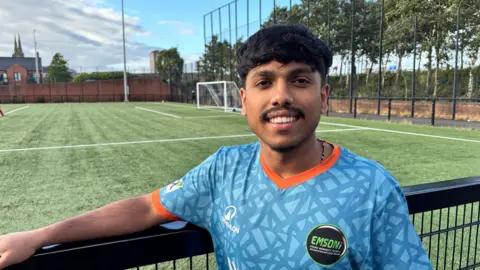 Nihal Shaji, a man with short, black hair and a thin moustache, smiles at the camera while leaning one arm on a black railing which overlooks a football pitch on a sunny day. He is wearing a light blue football shirt with an orange collar and sleeve edges. A black badge on his top has the abbreviation EMSONI in green lettering, which stands for Ethnic Minority Sports Organisation Northern Ireland.