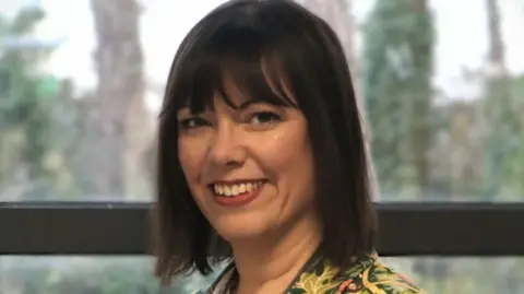 Corby Technical College Anna Jenkins is smiling at the camera. She is standing in front of a window and has dark brown hair in a bob and is wearing a green patterned jacket.