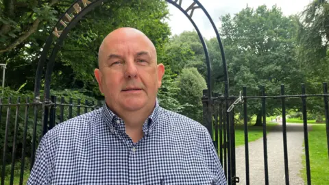 BBC A man is looking into the camera. He's standing in front of gates. he's wearing a checked shirt. Trees, grass and bushes are in the background. 