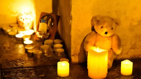 A white teddy bear sits against a cream wall with three candles in front of it. There are a collection of about a dozen tealights to the right of the teddy.