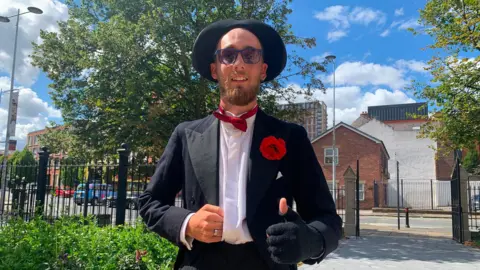 A man dressed in a hat and fingerless glove and bow tie shows the thumbs up sign