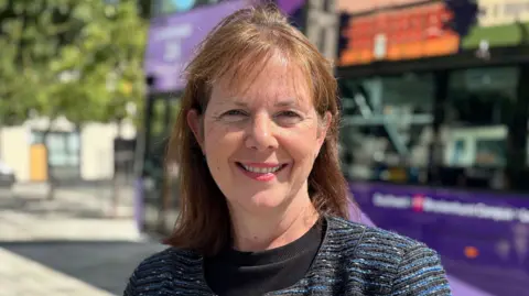 A head and shoulder image of a smiling woman standing in front of a bus