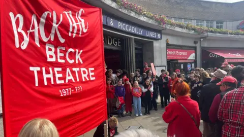 Tolu Adeoye crowds of people holding 'back the beck theatre' banners outside the entrance to uxbridge tube station