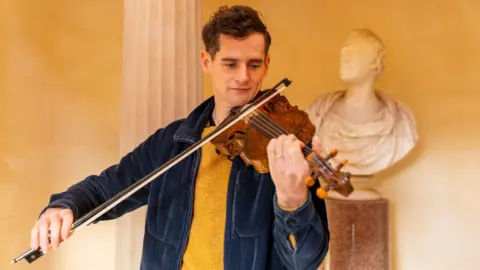 Ewen Handerson performing with the Gregg fiddle. He's wearing a yellow jumper and blue jacket, while playing the fiddle. A white marble bust is behind him.