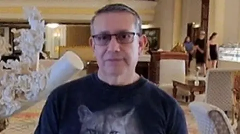 Jorge Ortega wearing glasses and a black T-shirt with a cat on, seemingly in the lobby of a hotel with guests over his left shoulder