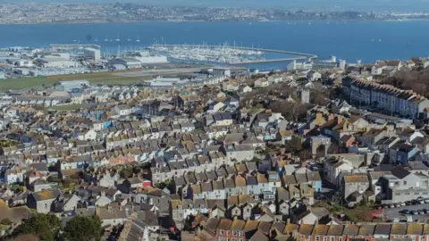 Aerial view across Portland looking towards Weymouth. In the foreground, taking up most of the photo, is the town of Fortuneswell, consisting of mostly terraced houses built on a hill. Beyond the houses is Portland Port and Portland Habour and a large marina full of small boats. On the other side of the harbour is the Wyke Regis area of Weymouth.