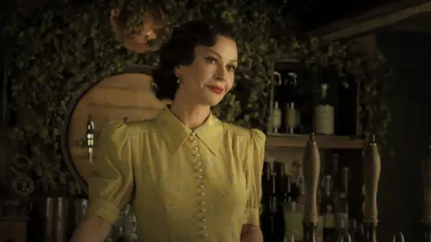 Catherine Zeta Jones in a yellow dress. She has short dark hair and is standing behind an old style bar. 