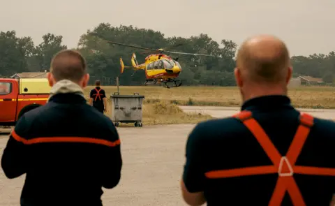 Laurent Perpigna / EPA Two firefighters with their backs to the camera as they look out over a scene of a helicopter taking off on tarmac in south-western France on Monday.