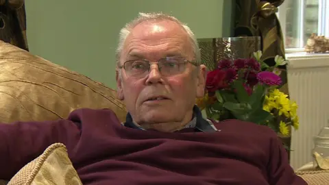 Alan Knight sits on his sofa giving a TV interview. He has light grey hair and glasses.