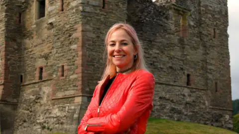 BBC/Rare TV Prof Alice Roberts standing in front of the ruined castle keep. She has pink tinted hair and is wearing a red jacket. Her arms are folded and she is smiling broadly. 