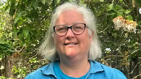 Charlotte Clark smiles at the camera while standing outside. She has short grey hair and wears glasses with a light blue top.