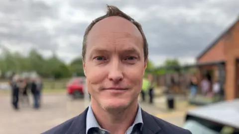 Alex Dunlop/BBC Scott Hussey, a Reform UK councillor, is standing outdoors in a car park. He is wearing a light grey shirt and blazer. He is looking directly at the camera and smiling.