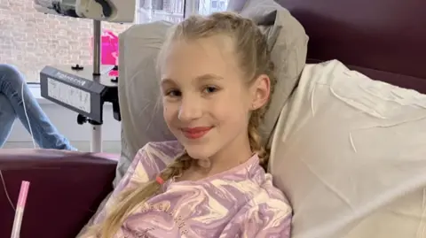 Family photo A girl with blonde hair in a plait, wearing purple and white pyjamas, sitting up in a hospital bed.
