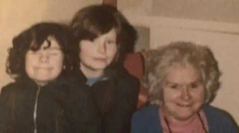 Diane Barnes An old photograph of two young girls with dark hair and their grandmother. The girls are standing together next to an older woman who's sitting in a chair. They're all looking at the camera. 