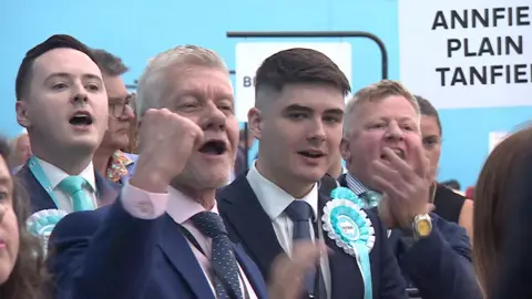 BBC Reform UK celebrating. There are four men pictured, two are wearing rosettes.