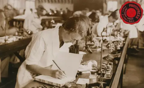UCL Black and white photo shows a woman in science lab writing in a large book. Others are behind her using equipment
