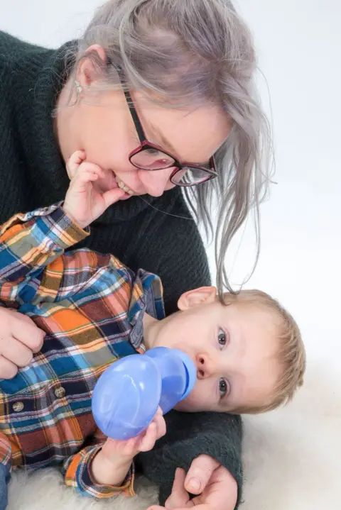 Family handout A woman with glasses and dyed grey hair is smiling looking down at a toddler in her arms who is drinking water from a toddler bottle. The little boy is wearing a checked orange and blue shirt, and it looks like a professional photograph. 