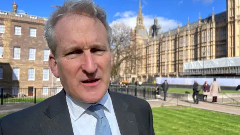 MP Damian Hinds stands outside Parliament during an interview with the BBC. He is wearing a dark grey suit and a light blue shirt and tie.