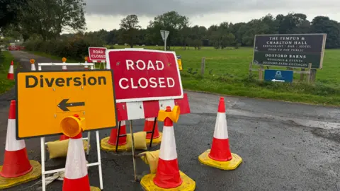 Katryna Shell  A road closure sign is at the heart of the picture with a diversion sign next to it and a number of traffic cones. A second road closure sign can be seen behind.