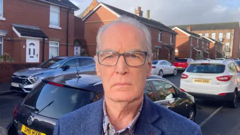 Gerry Kelly, a man with short, grey hair and glasses, stands on a residential street looking at the camera. He is wearing a checked shirt and a blue tweed-type blazer. There are parked cars and red bricked houses in the background. 