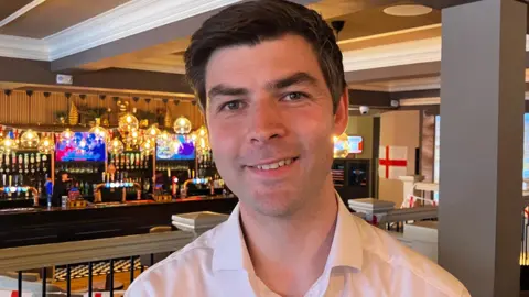 BBC News A man in a white shirt stands in front of a bar. Bottles and pendant lights can be seen in the background. 