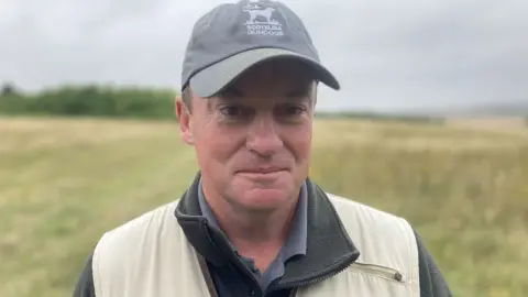 Mark Witham standing on Salisbury Plain wearing a baseball cap with a whistle around his neck, sat next to him is one of his five dogs, a golden Labrador
