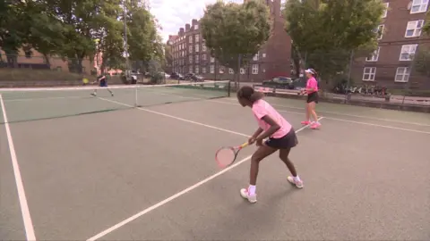 Young black girl wearing a pink top and black shorts on a tennis court ready to receive a ball