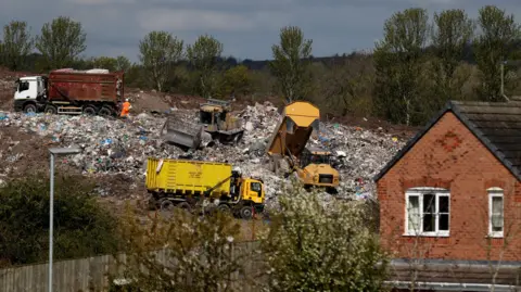 Reuters Three trucks emptying waste at a landfill site on a hillside behind a house.