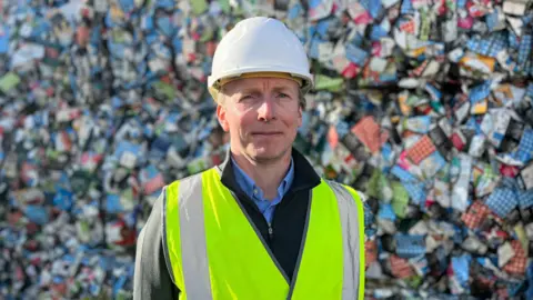 Michael Grime, Managing Director of Mayside Recycling, stood in a yellow high vis jacket, and white hard hat. He is wearing a navy quarter-zip jumper and a blue shirt. He is in focus and in the sunlight. He looks in his 40s, has blue eyes and is smirking. In the background, there are bails of milk cartons piled up behind him. 