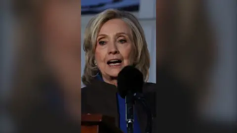 Former US Secretary of State Hillary Clinton wears a Royal blue shirt with a grey coat over that. She is standing behind a microphone.