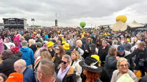 Festival crowd stretching into the background. Two hot air balloons can be seen at ground level in the background.