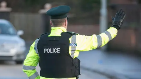 PA Media A police officer holds up their arm as they stop traffic on the street. He is pictured from behind and is wearing a police hat and vest, as well as a reflective coat. A car in the background is blurred.